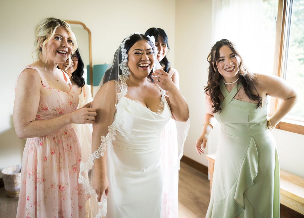 Bride in white gown laughing with bridesmaids in pink floral and sage green dresses during wedding preparations.