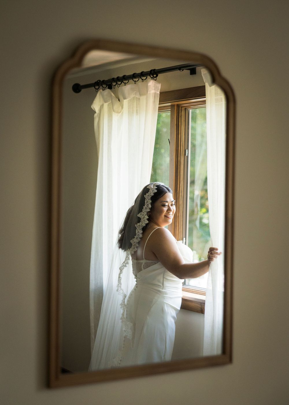 Bride in white wedding dress and veil admiring herself in an ornate gold mirror by a sunlit window.