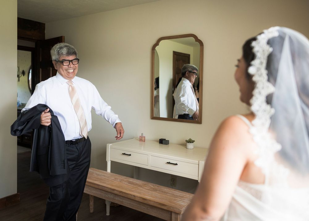 Bride in white veil faces groom in white shirt and black suit jacket in a warmly lit room with a mirror.