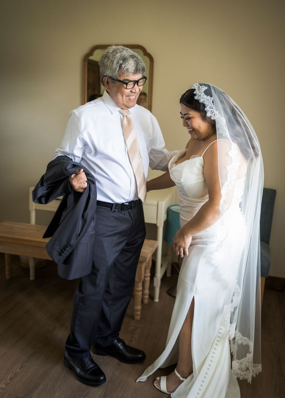 Bride in white gown and veil helps older man in glasses put on suit jacket indoors before wedding ceremony.