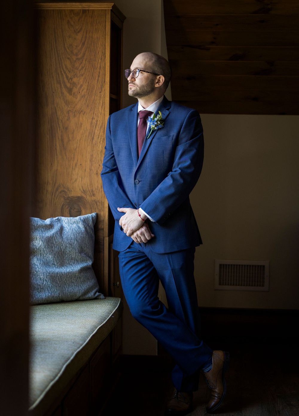 Dapper groom in navy blue suit with burgundy tie poses by window on wedding day.