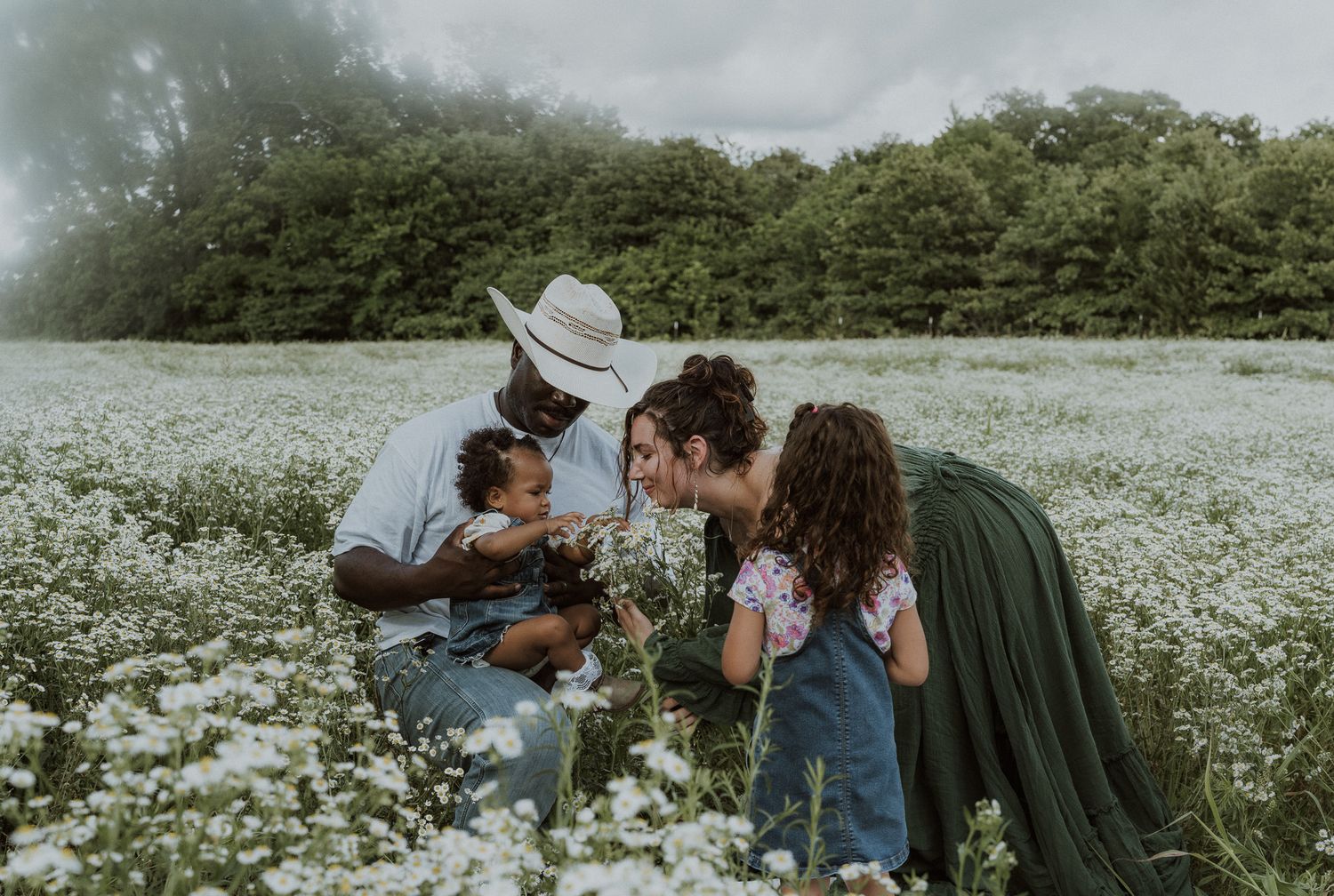 Family sharing a tender moment in a field of white wildflowers, with trees in the background.