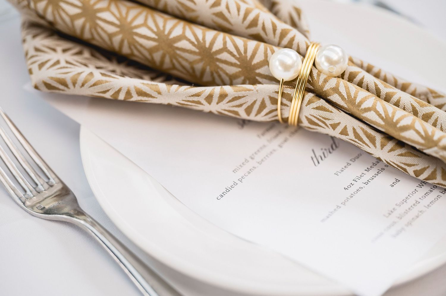 Gold geometric napkin with pearl napkin ring beside a fork and menu card on white plate.