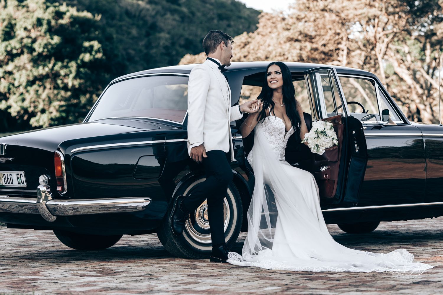 Elegant bride and groom posing beside a classic vintage black luxury car outdoors.