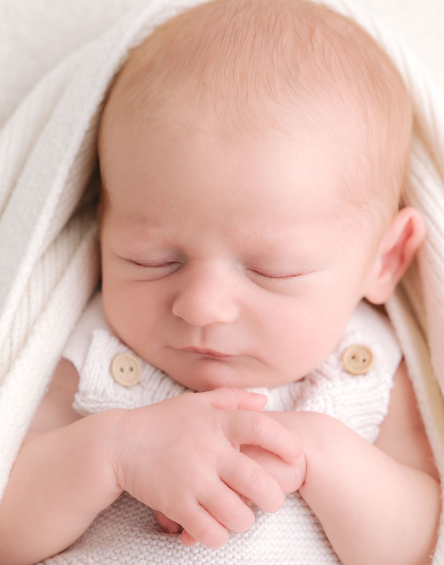 Peaceful newborn baby sleeping with hands clasped, wearing a white knit outfit, wrapped in a soft cream blanket.