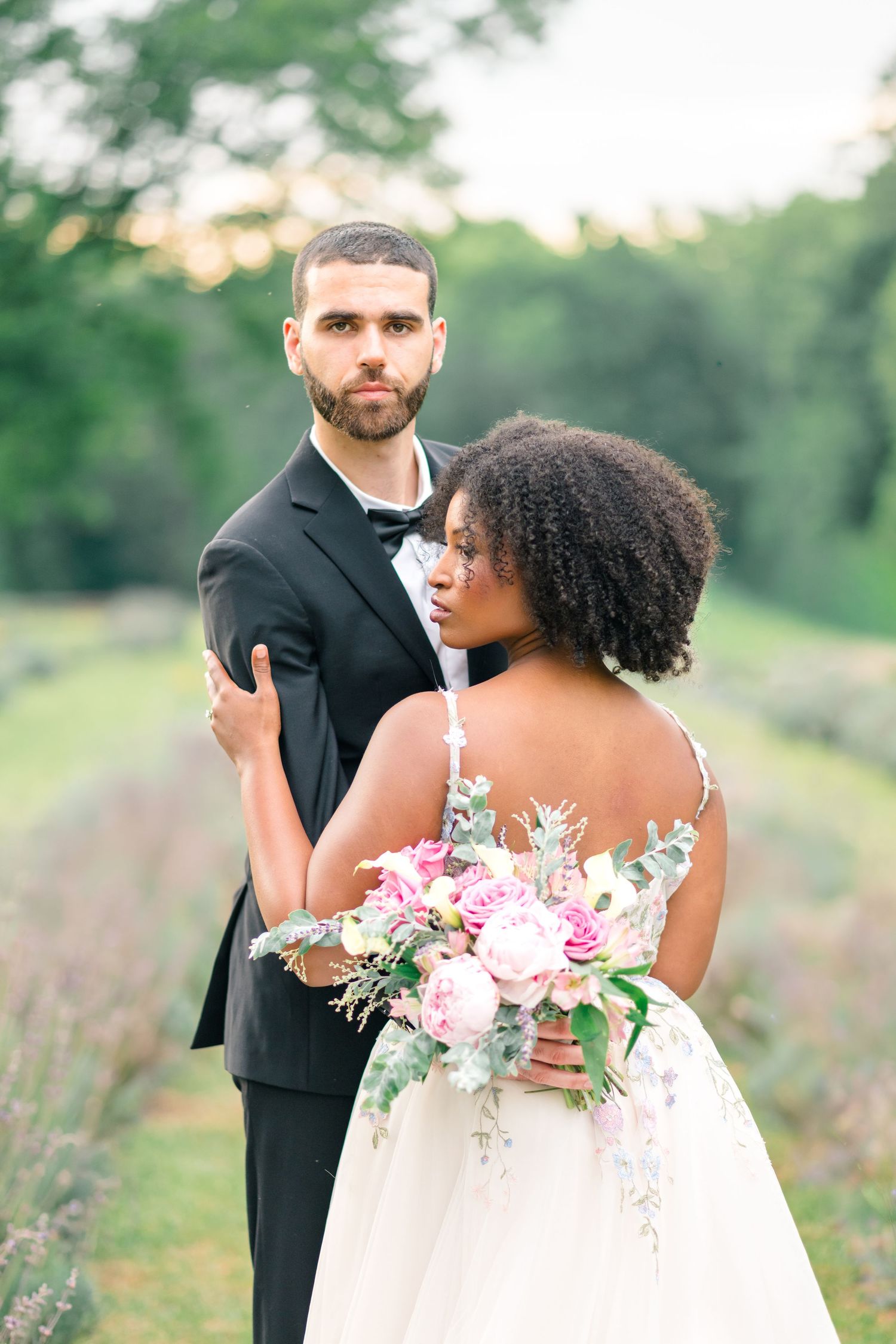 Interracial couple in wedding attire pose in lavender field, bride holding pink bouquet in white gown, groom in black tuxedo.