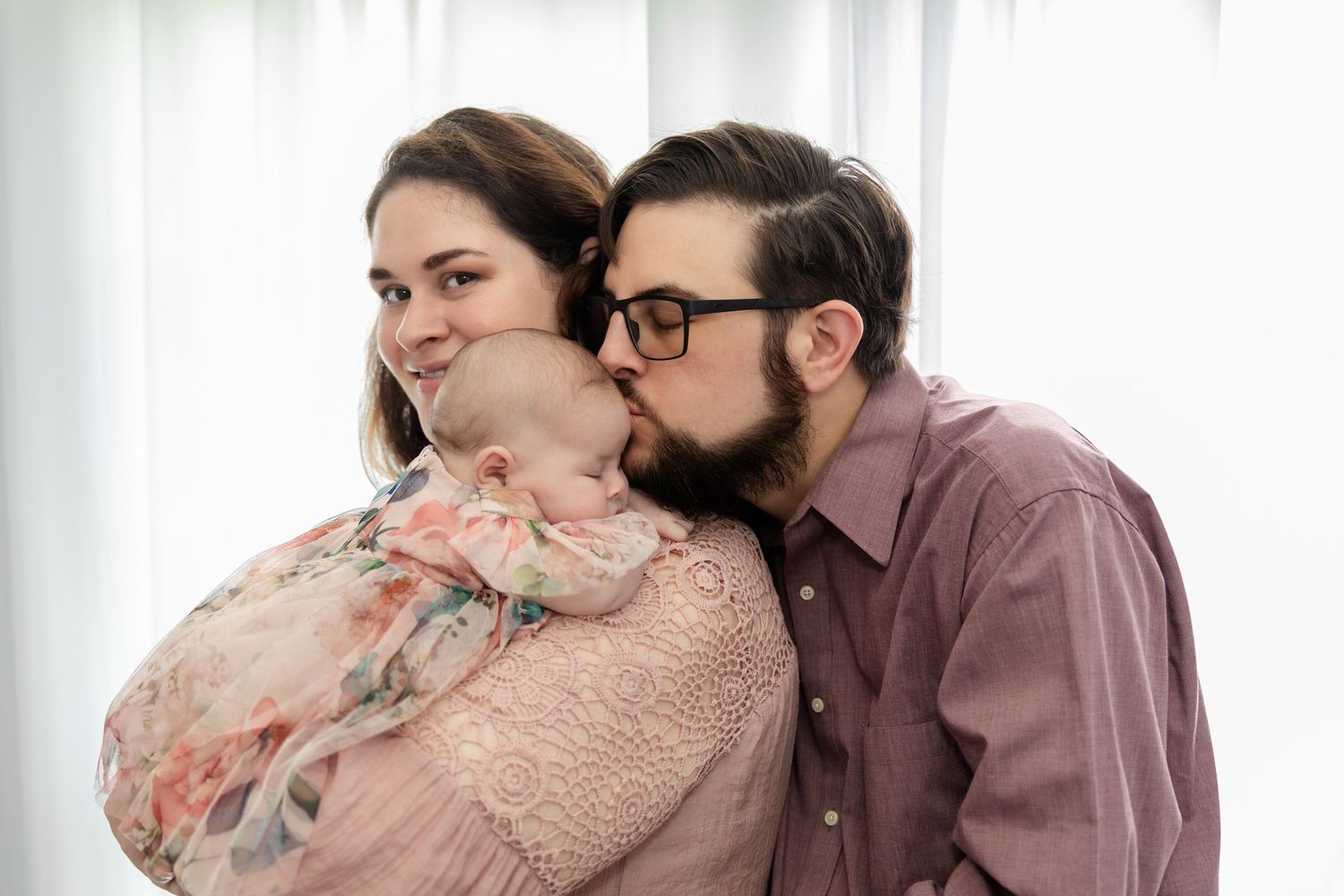New parents tenderly kissing their newborn baby wrapped in a floral blanket near a bright window.