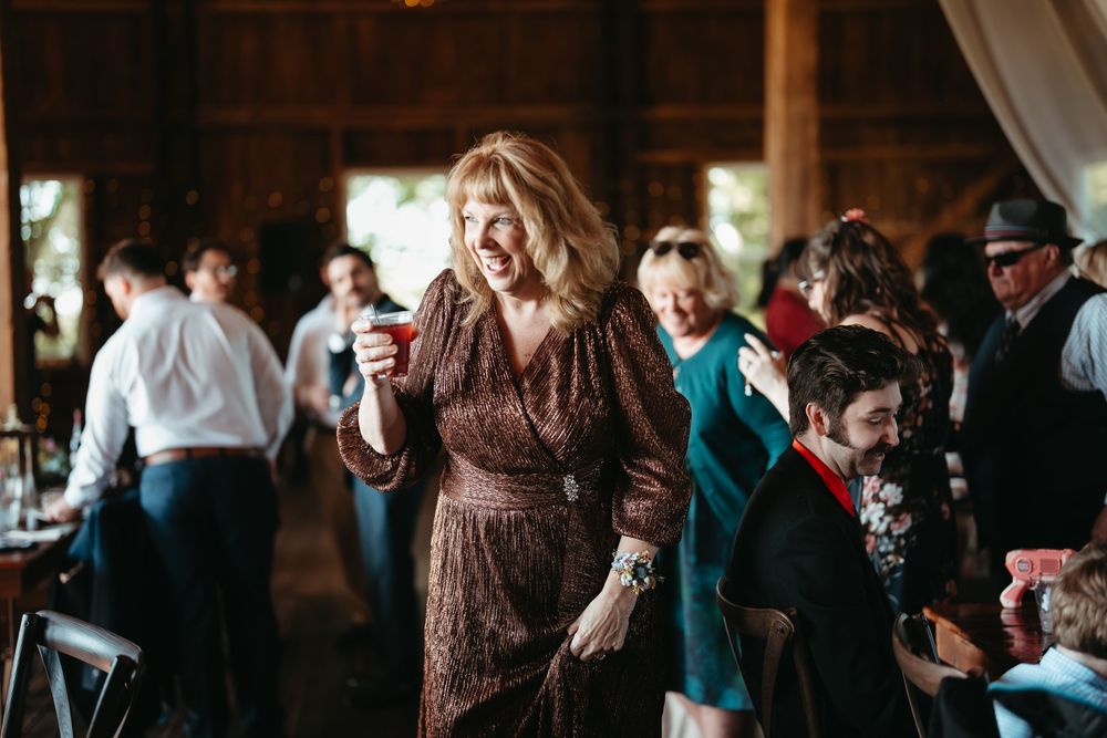 A woman in a brown wrap dress laughs while holding a drink at a rustic barn wedding reception.