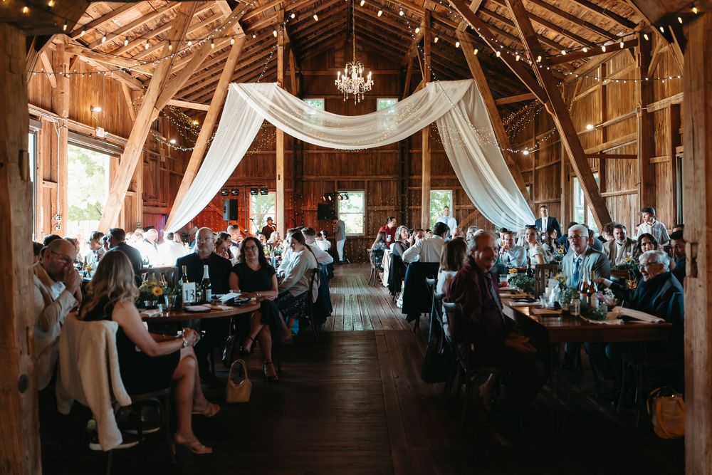 Rustic barn wedding reception with string lights, white draping, wooden beams, and guests seated at long tables.