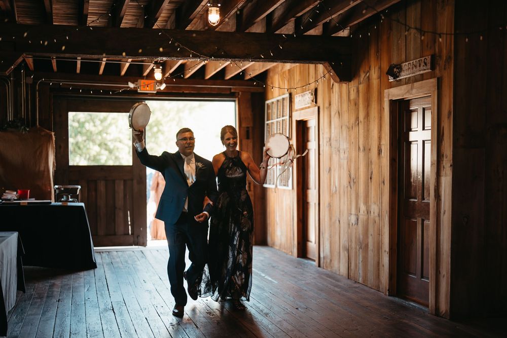 Couple makes grand entrance into rustic barn wedding reception venue with string lights and wooden walls.