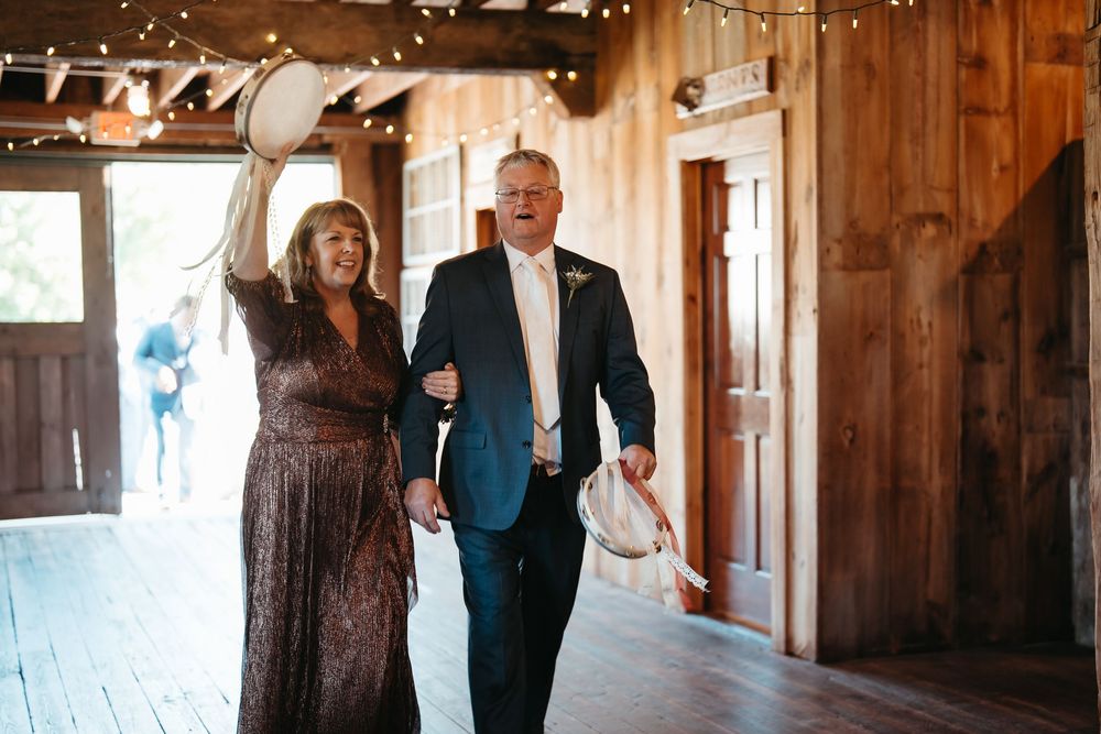 Couple making a joyful grand entrance into a rustic barn wedding reception with string lights.