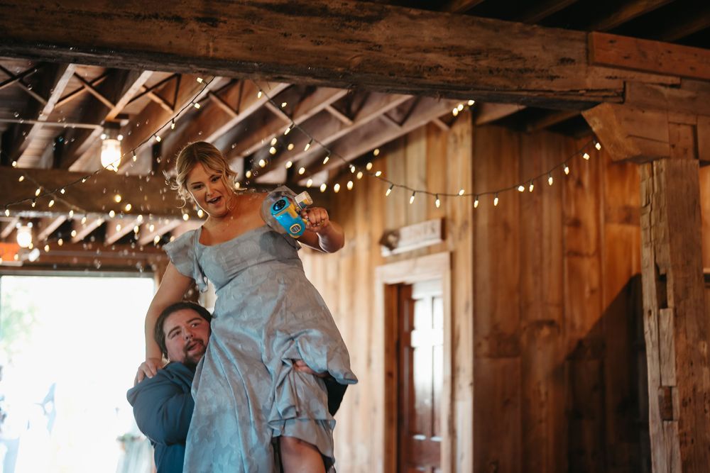 Joyful wedding guest lifted on shoulders in decorated rustic barn venue with string lights.