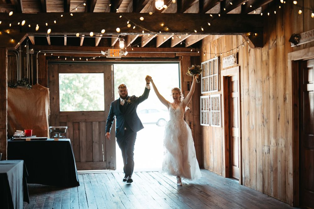 Bride and groom make grand entrance into rustic barn reception with string lights overhead.