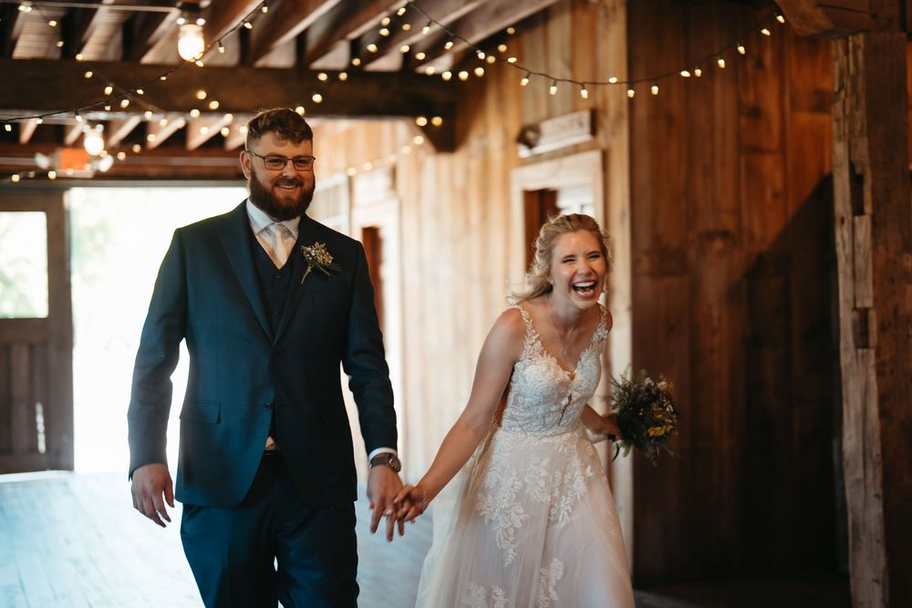 Bride and groom walk hand in hand laughing through rustic barn venue adorned with string lights on their wedding day.