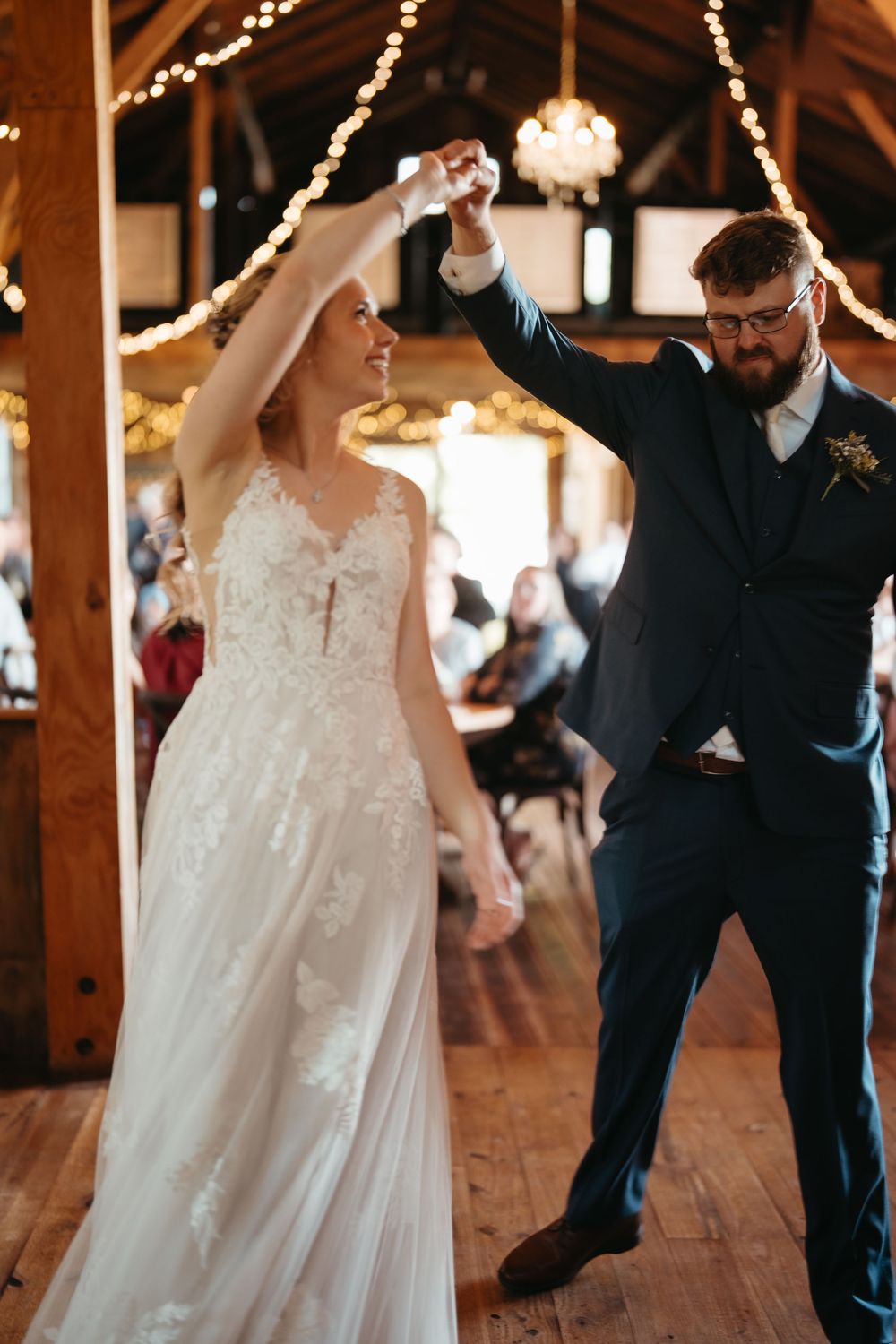 Bride and groom share first dance in warmly lit rustic barn wedding venue with guests.