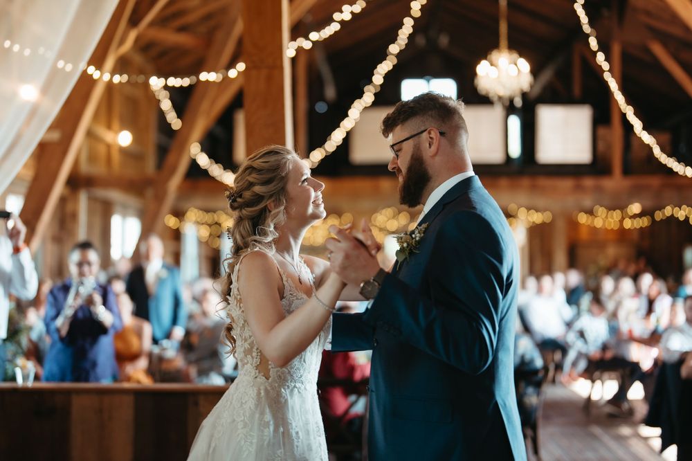 Bride and groom share their first dance in a rustic barn venue decorated with warm string lights.