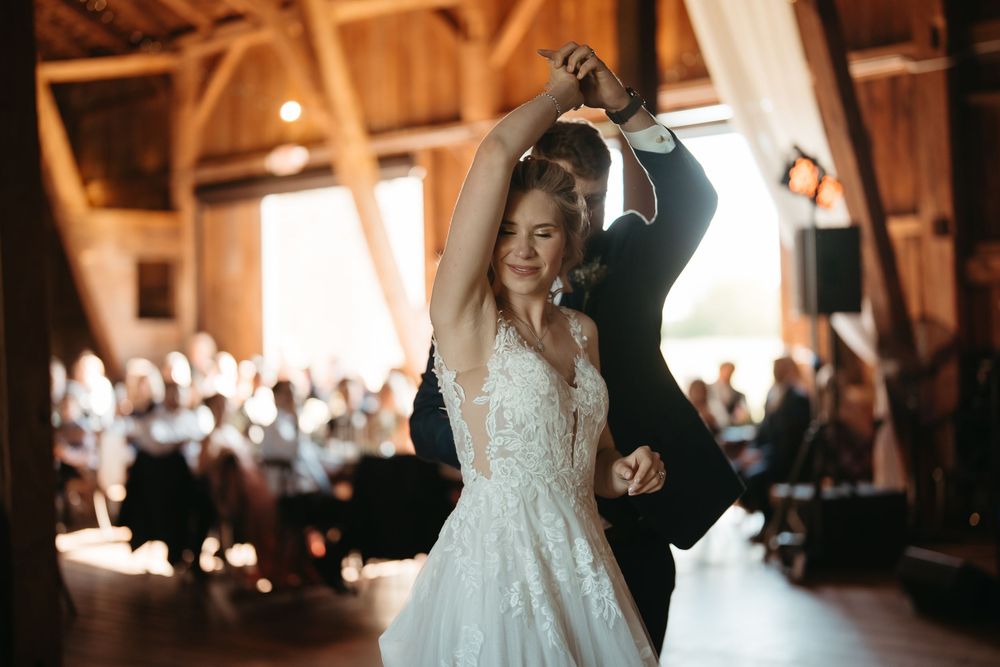 Bride twirls in her lace wedding gown during first dance in a warmly lit rustic barn reception.