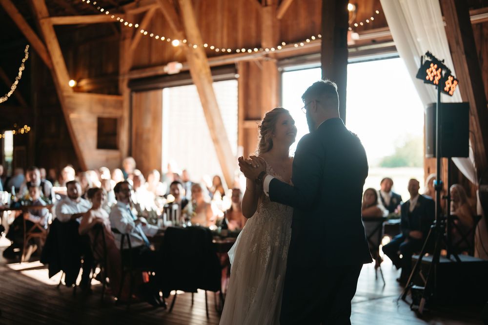Bride and groom share their first dance in a warmly lit rustic barn surrounded by wedding guests.