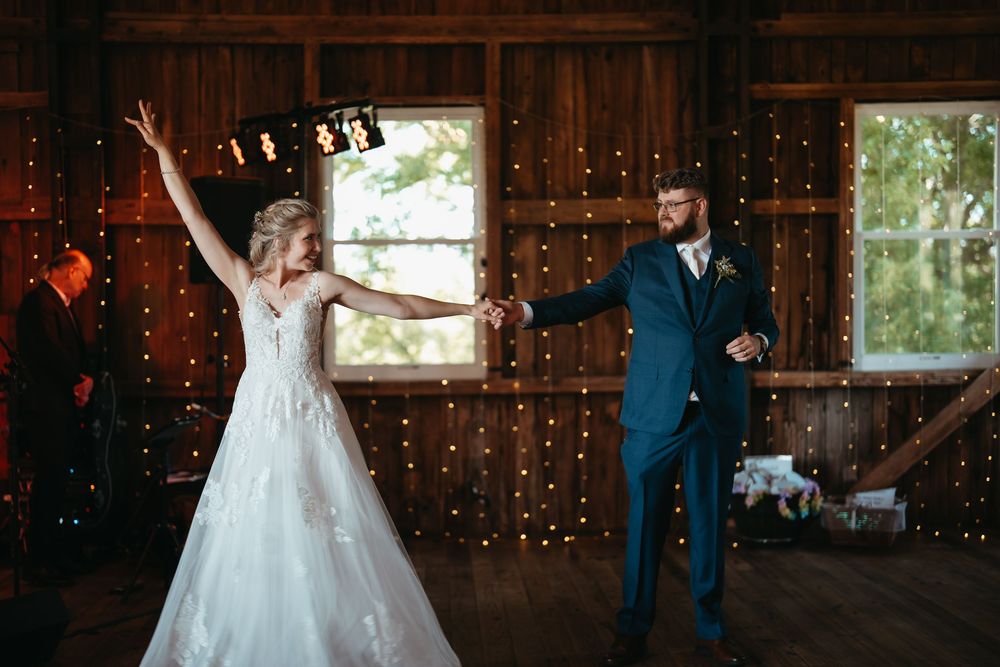 Bride raises her arm joyfully while dancing with groom in a rustic barn wedding venue with fairy lights.