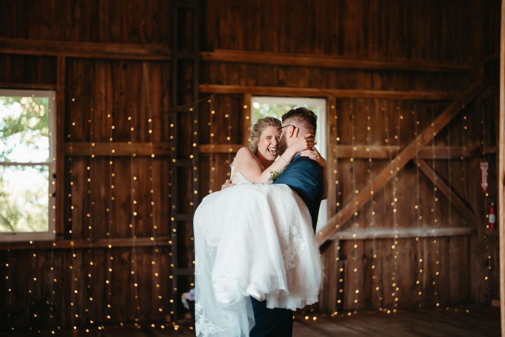 Groom lifting smiling bride in rustic barn wedding venue adorned with twinkling string lights.