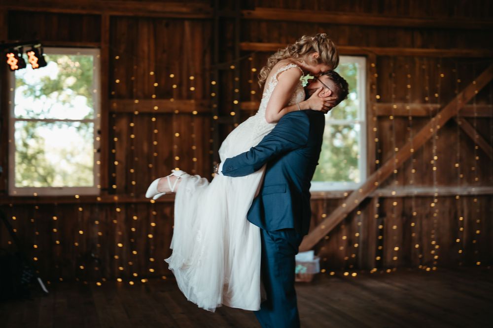 Groom lifts bride in a romantic moment surrounded by twinkling fairy lights in a barn.