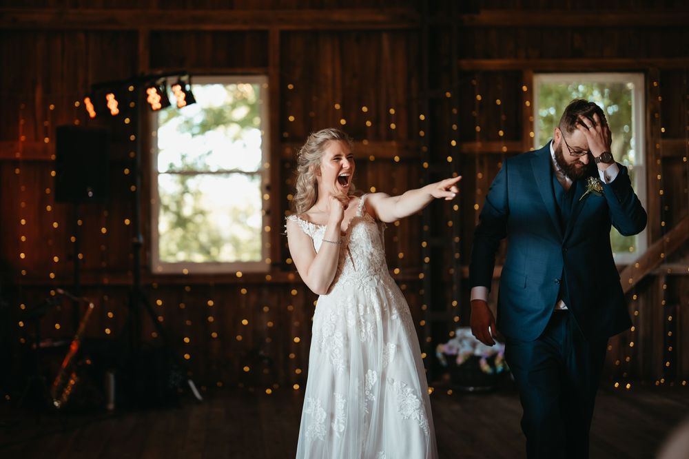 Bride laughs and points while groom reacts with surprise during fun wedding reception moment.