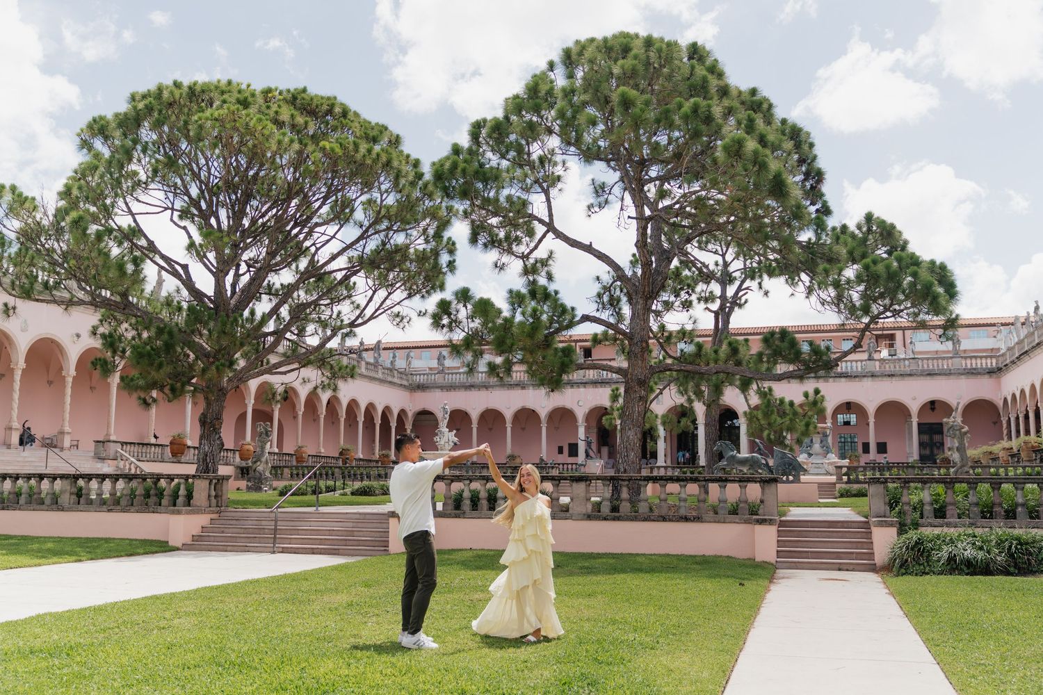 Couple dancing on green lawn at Ringling Museum courtyard with pink arched building and tall pine trees backdrop.