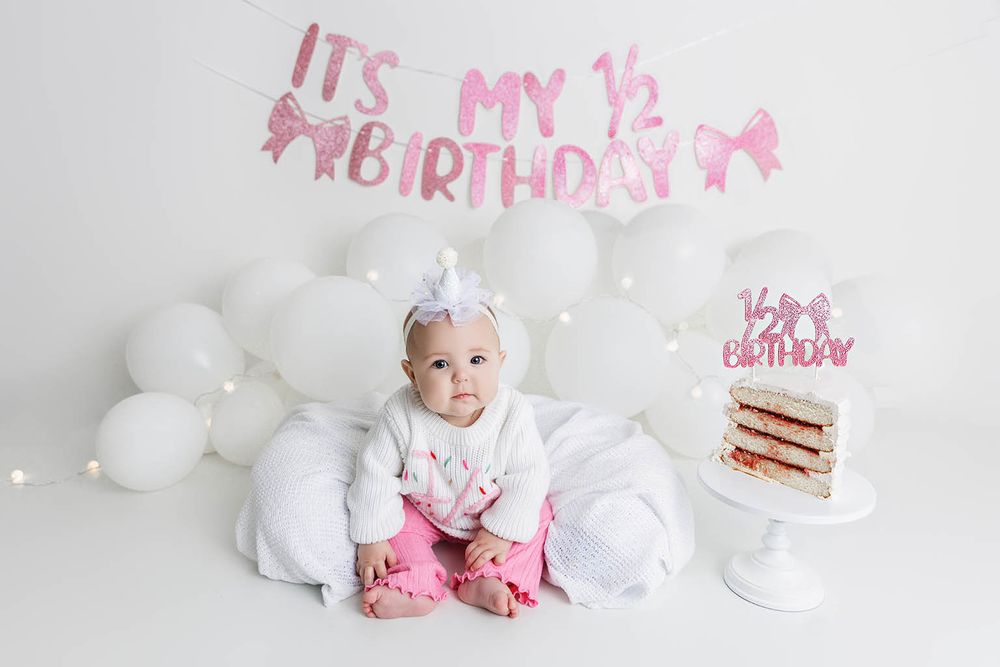 Baby girl in pink and white outfit celebrates her half birthday with balloons and cake.