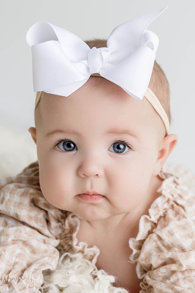 Adorable baby with blue eyes wearing a large white bow headband and ruffled outfit.