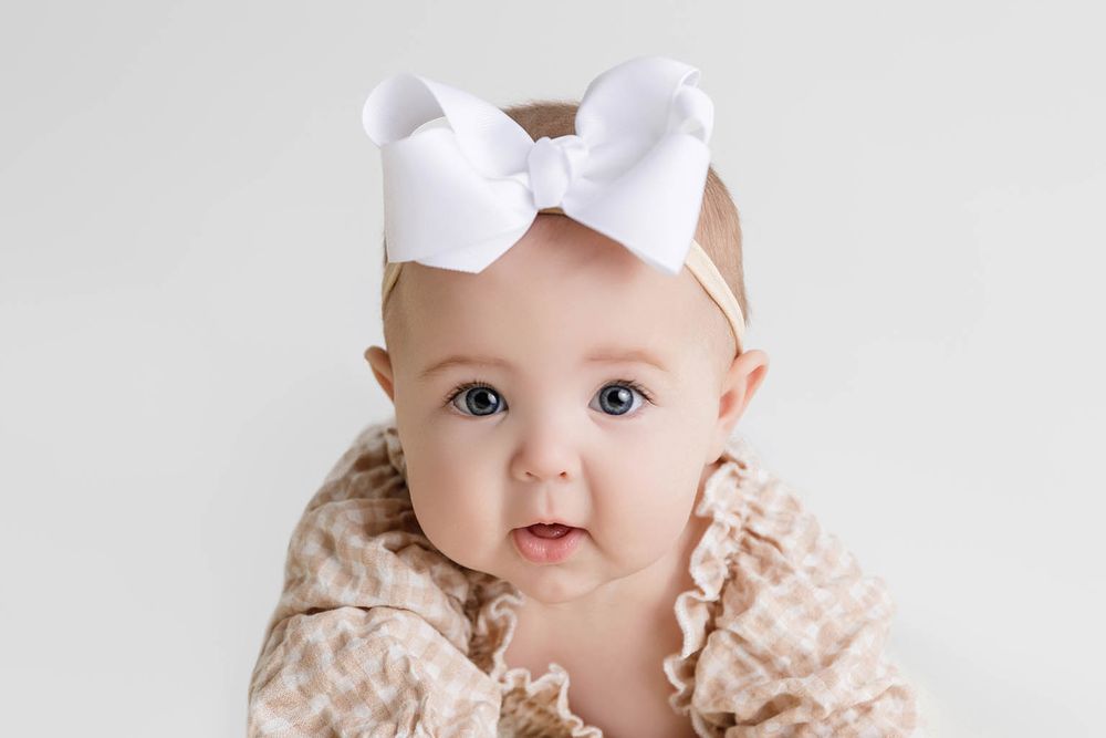 Adorable baby girl wearing a white bow headband and ruffled floral outfit, looking at camera.