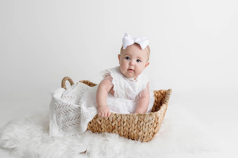 Baby girl in white lace romper and bow headband sitting in woven basket on fluffy white rug.