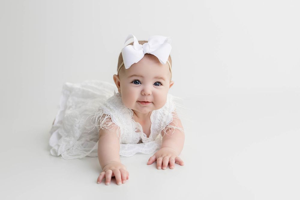 Adorable baby girl in white lace outfit and bow headband doing tummy time on white background.