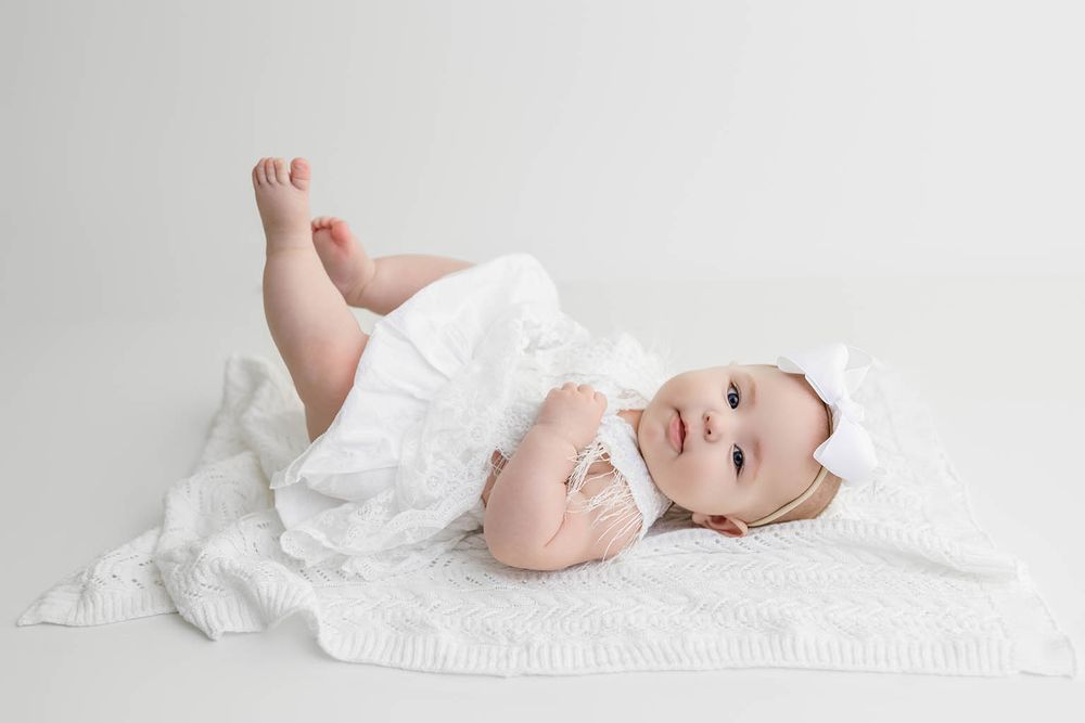 A baby in a white dress and bow headband lies on a white blanket, looking at the camera in a studio setting.