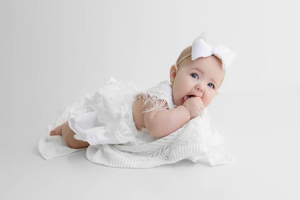 Sweet baby girl in white lace romper and bow headband lying on soft blanket, fingers in mouth.