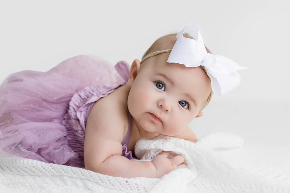 A cute baby girl in a purple tutu dress and white bow headband lying on a white blanket.