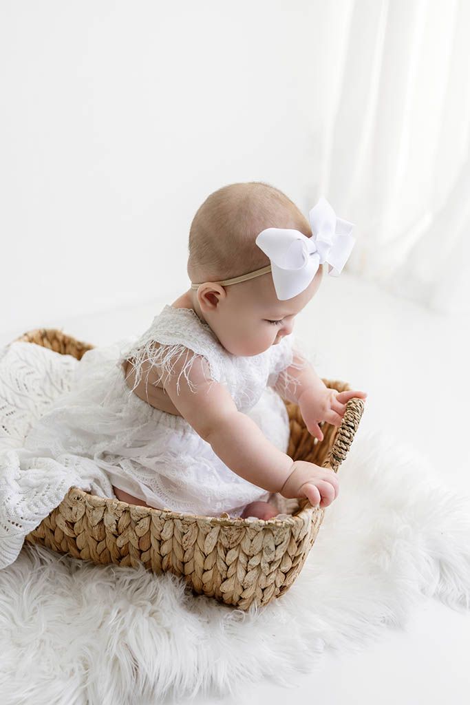 Cute baby girl in white lace dress with bow headband sitting inside a woven basket on fluffy rug.
