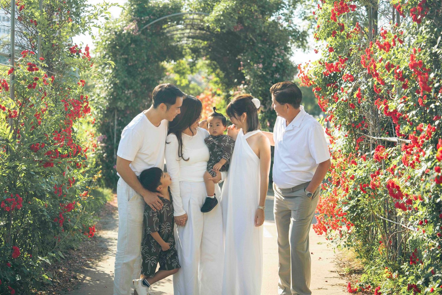 Family gathering in a lush garden archway adorned with vibrant red flowers, dressed in elegant white outfits.