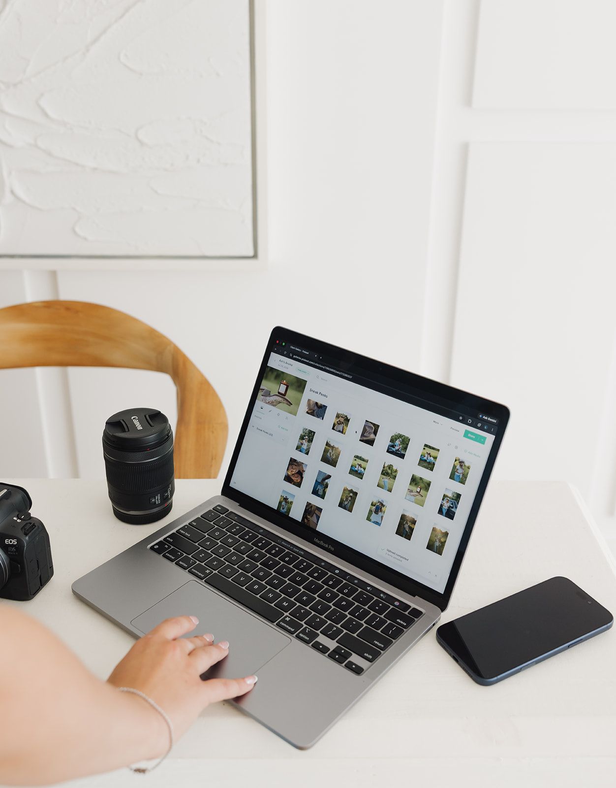 A person uses a MacBook laptop at a white desk with a Canon DSLR camera, lens, and smartphone nearby.