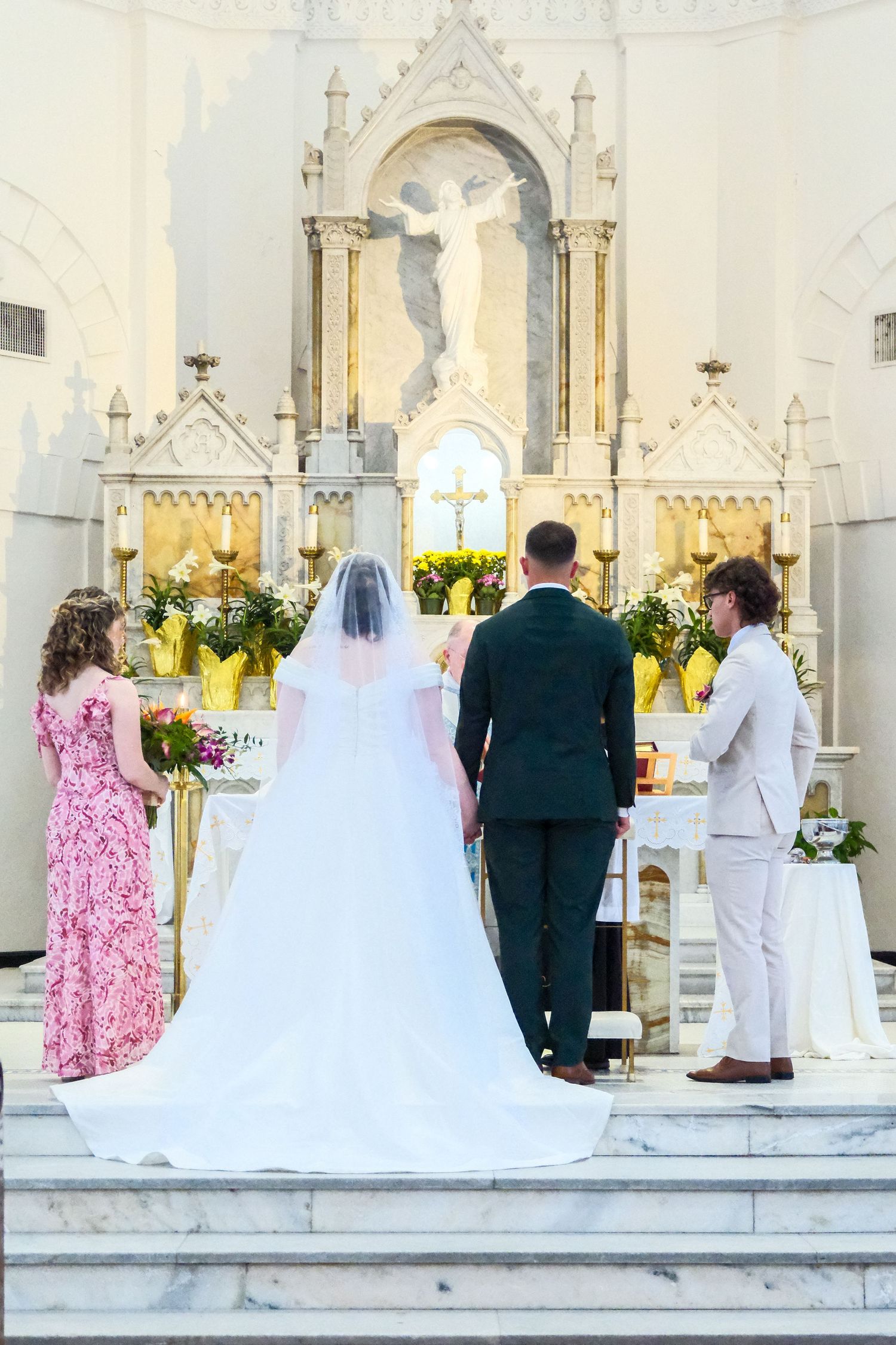 Bride in white gown with cathedral veil stands at ornate church altar with groom during ceremony.