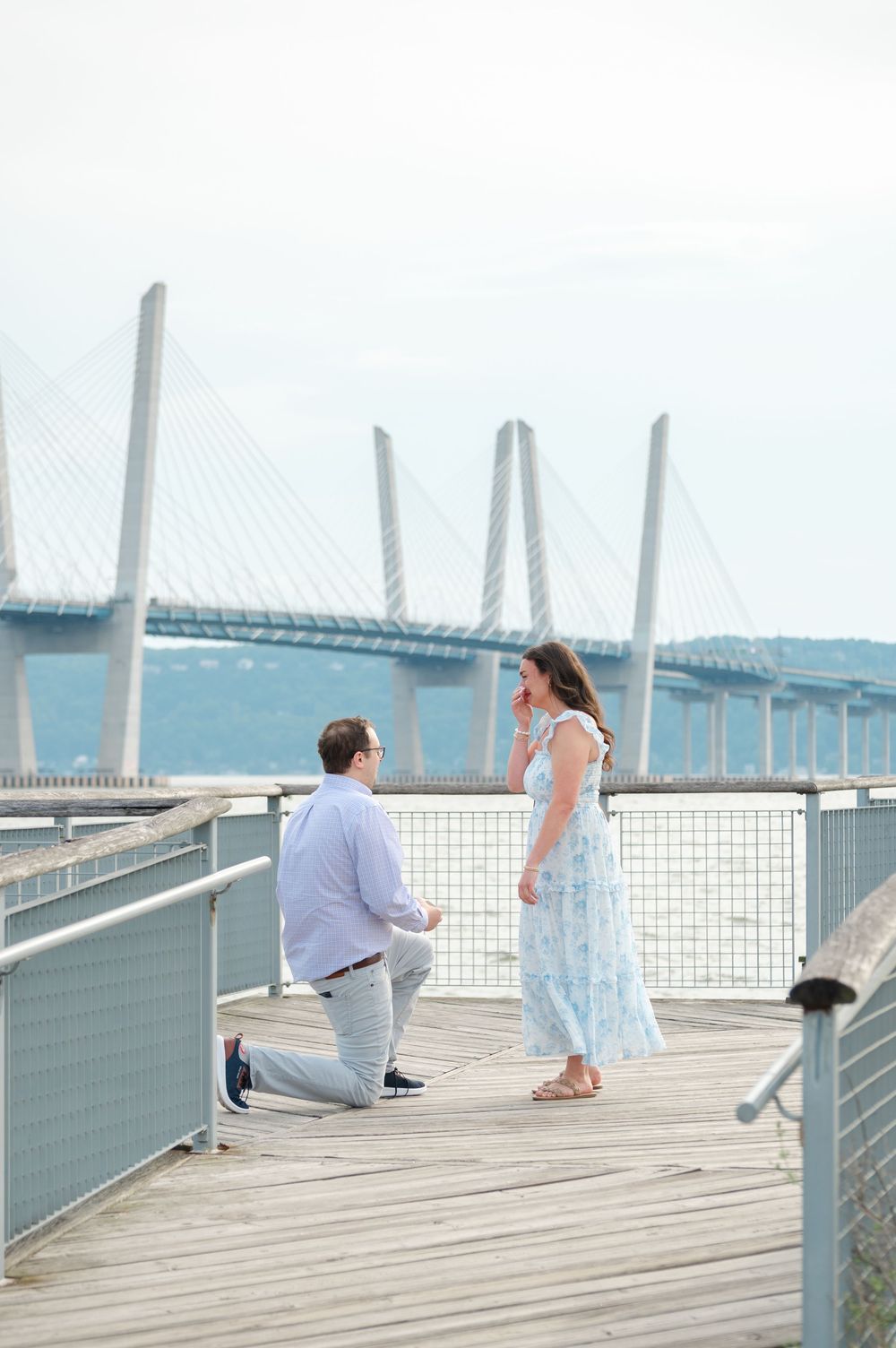 Man proposes on a wooden boardwalk with a cable-stayed bridge in the background as woman reacts with surprise.