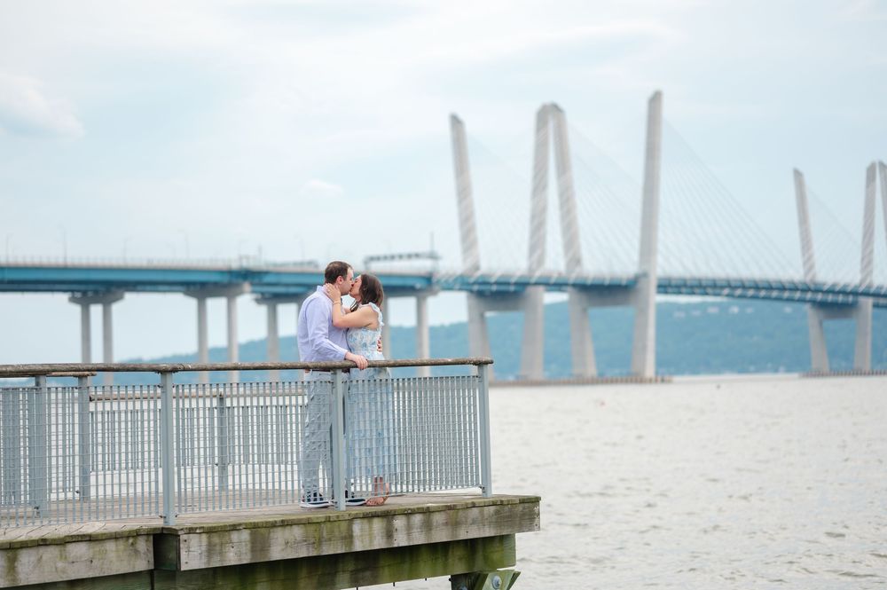 Couple kissing on a dock with a cable-stayed bridge spanning the river in the background.