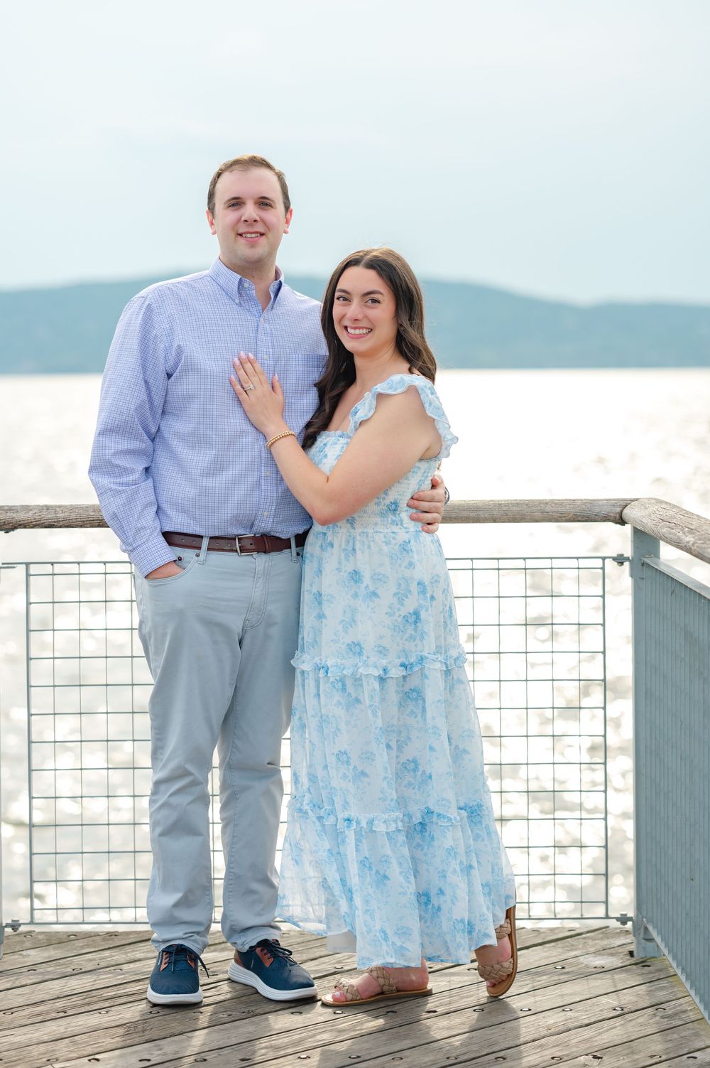 Smiling couple posing together on a wooden pier overlooking the water, woman in a blue floral maxi dress.
