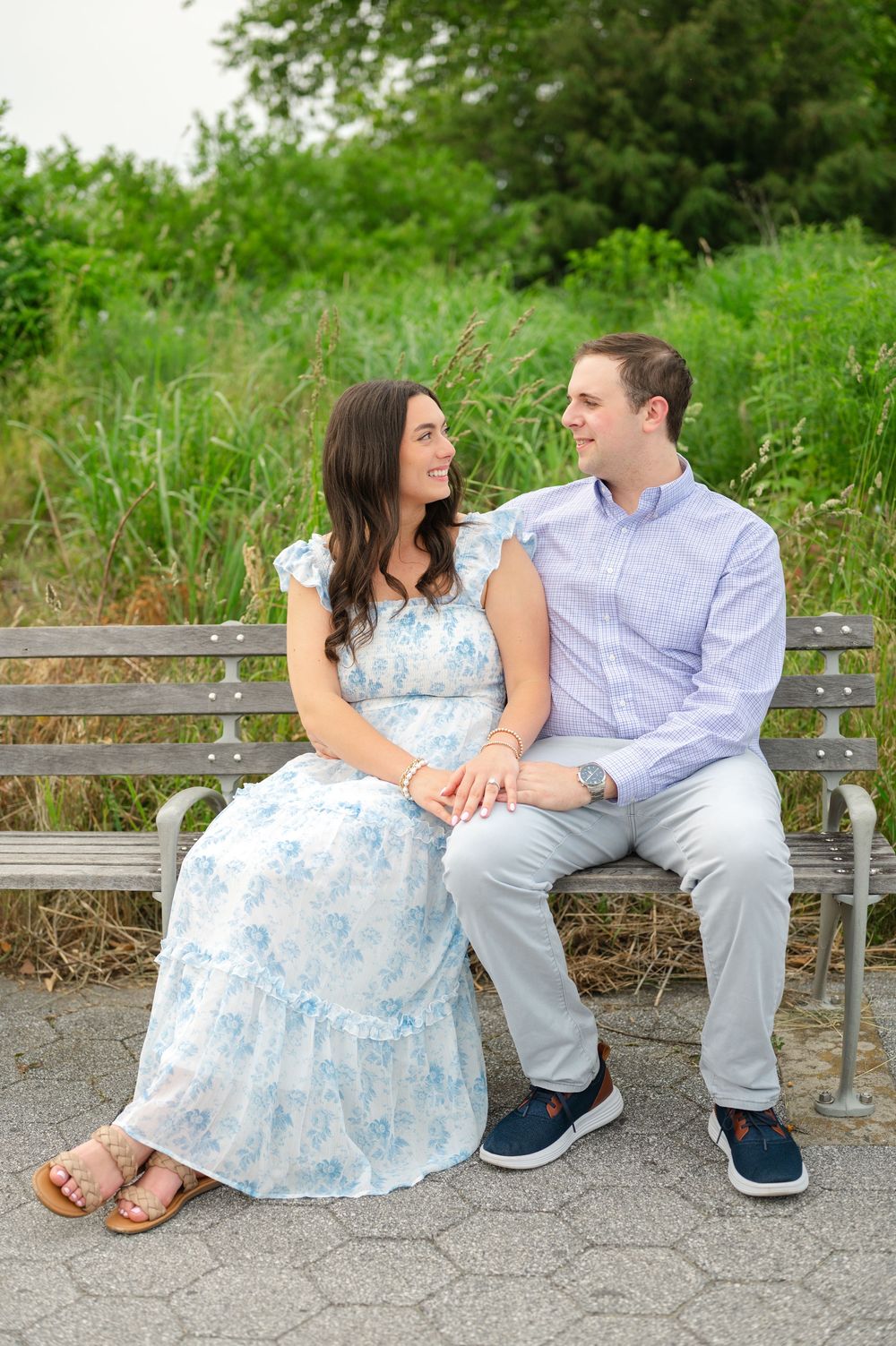 Engaged couple sitting on a park bench smiling at each other with lush greenery behind them.