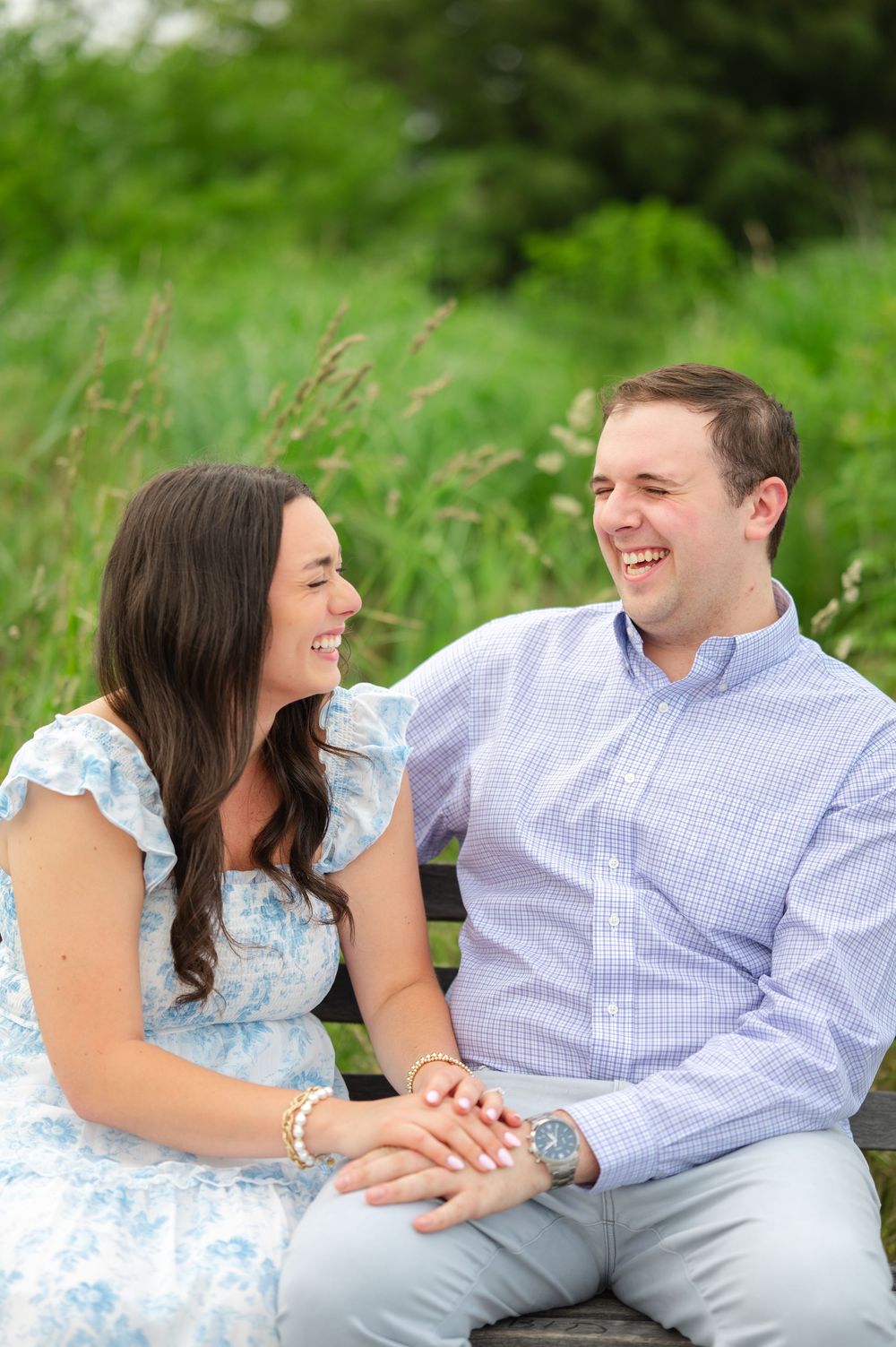 A happy couple sits on a bench laughing together outdoors, woman in blue floral dress, man in light blue checkered shirt.