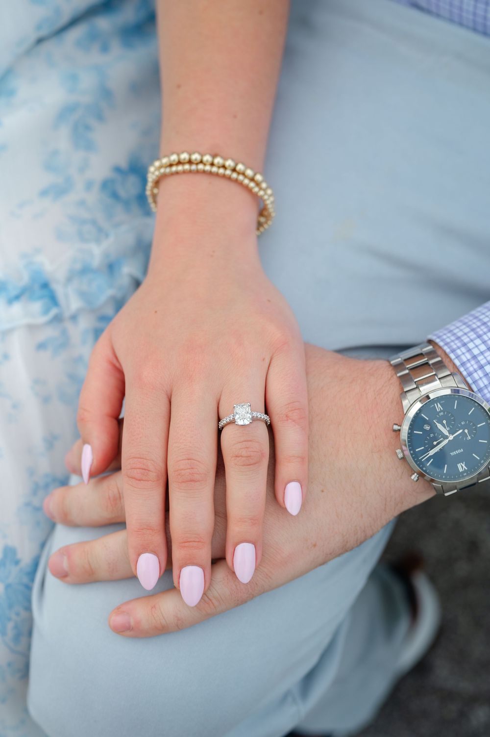 Close-up of an engagement ring on a woman's hand with pink nails resting on her fiance's lap.