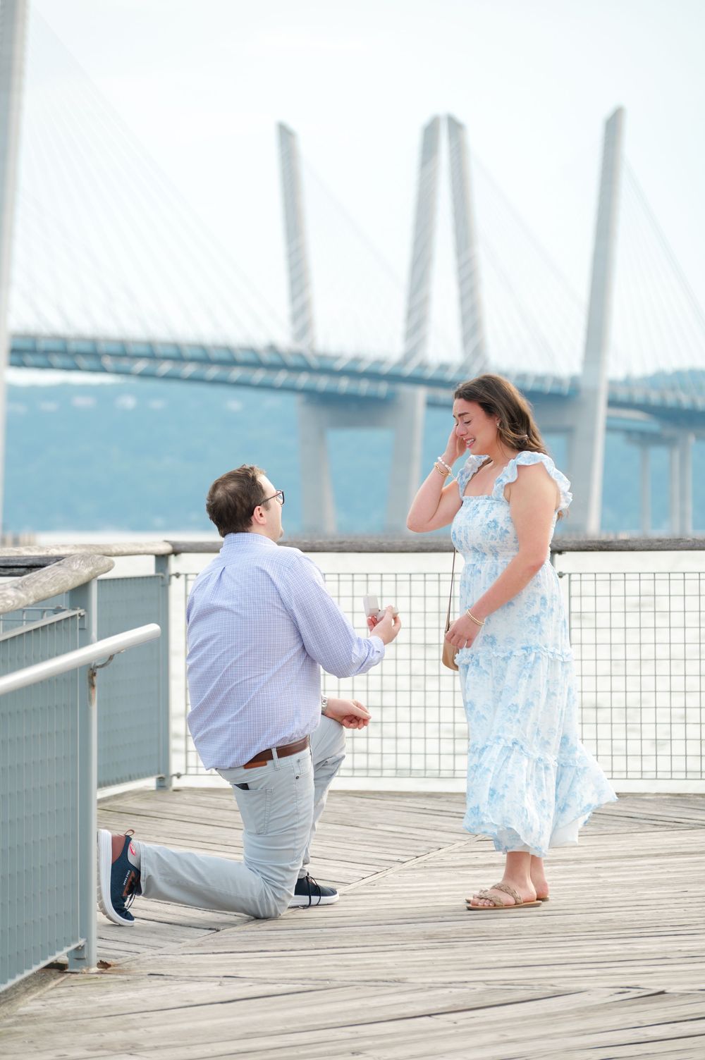 Man proposing on one knee to woman on a wooden pier with a cable-stayed bridge in the background.
