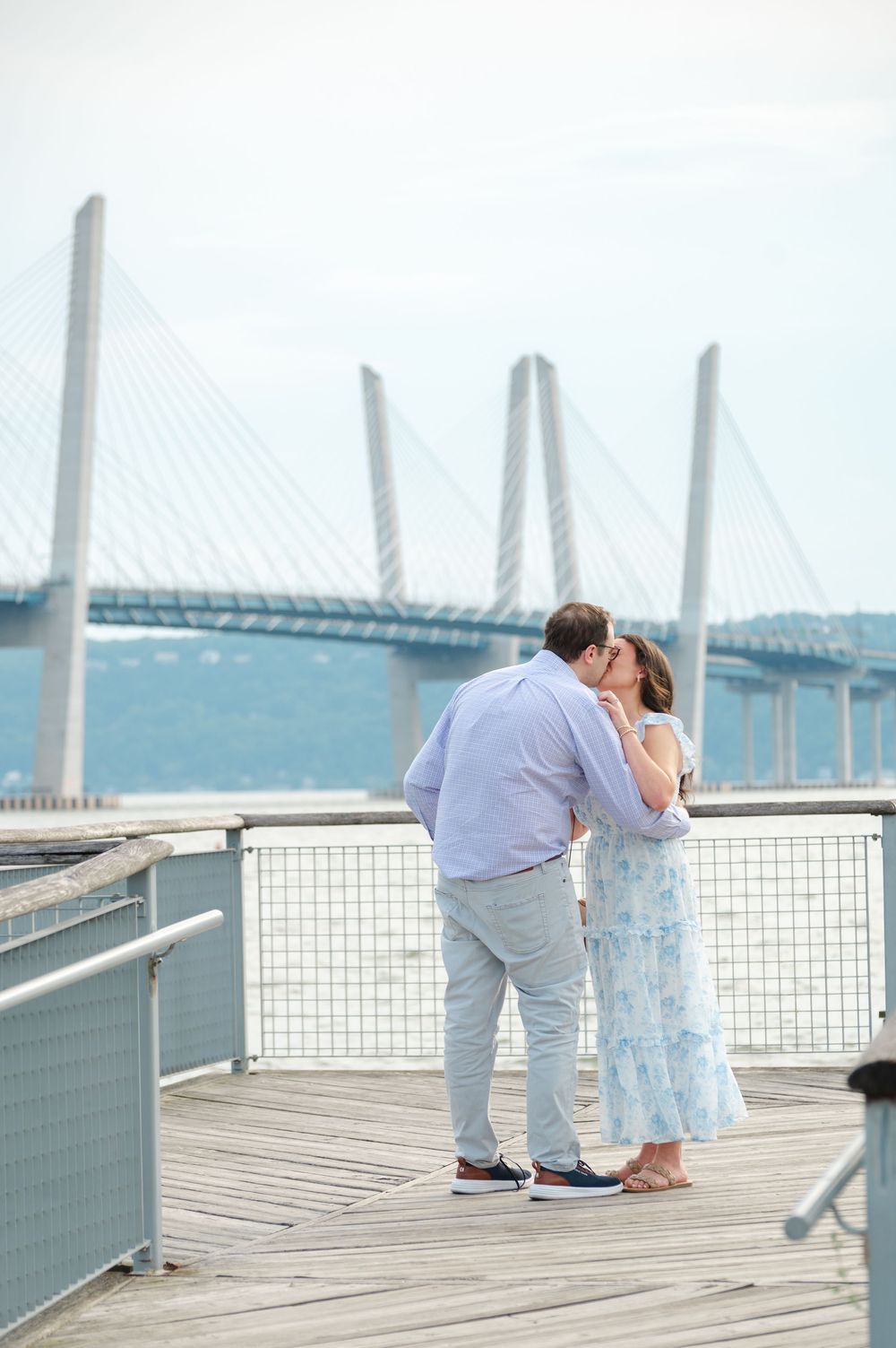Couple sharing a romantic kiss on a wooden pier with the Tappan Zee Bridge in the background.