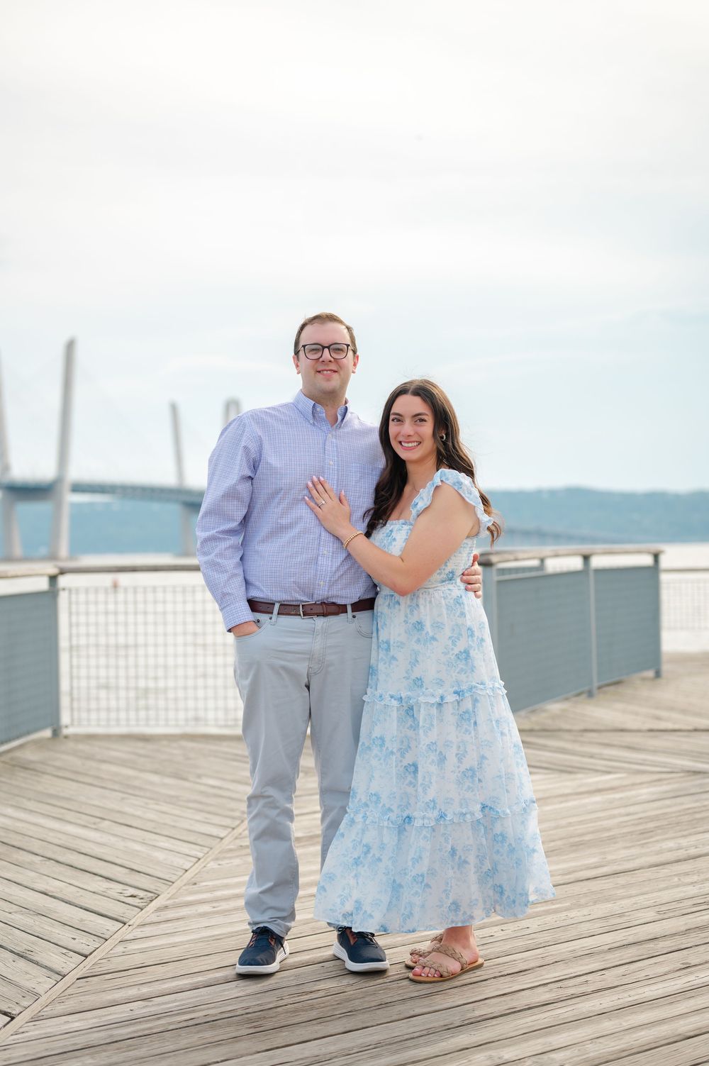 Couple smiling together on a waterfront boardwalk, woman in floral blue dress, man in blue shirt and gray pants.