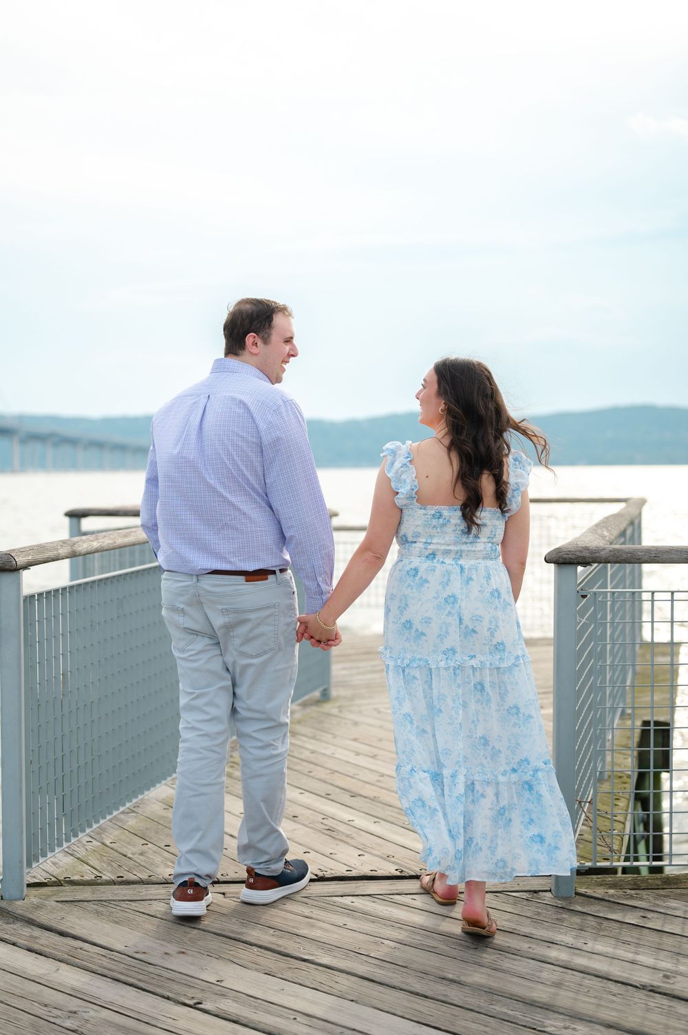 Couple holding hands on a waterfront pier, woman in blue floral dress and man in light blue shirt and gray pants.
