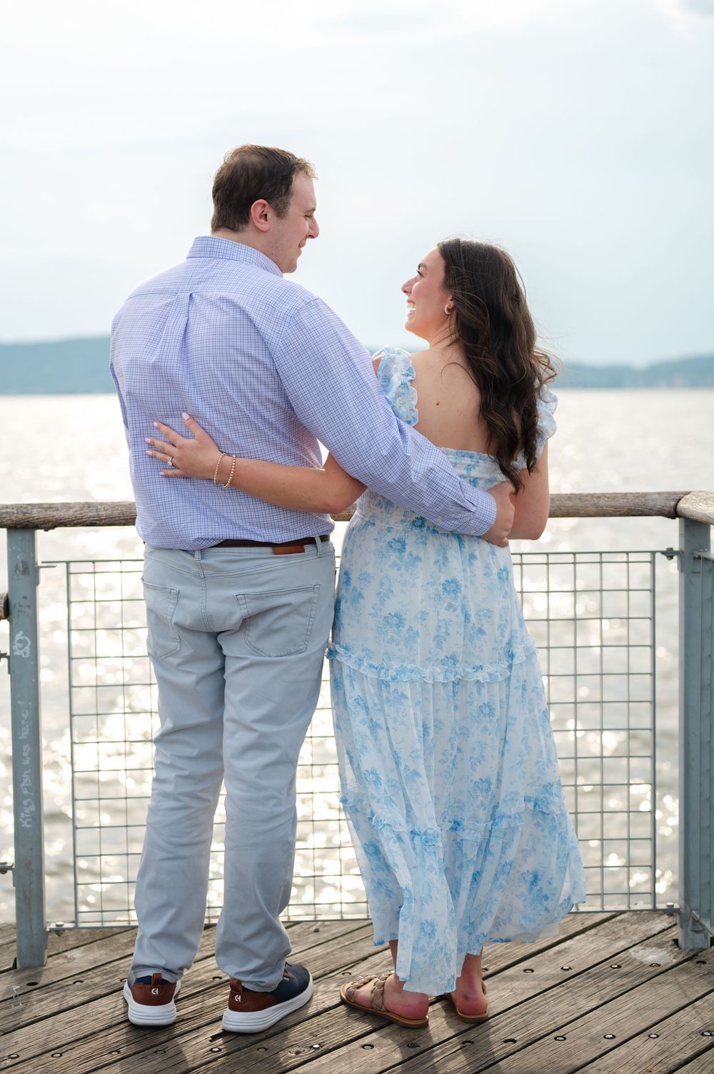 Couple embracing and gazing at each other on a waterfront pier with sparkling water behind them.